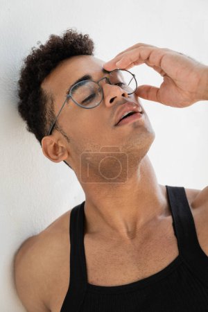 Portrait of relaxed and stylish young cuban man in black sleeveless t-shirt touching eyeglasses 