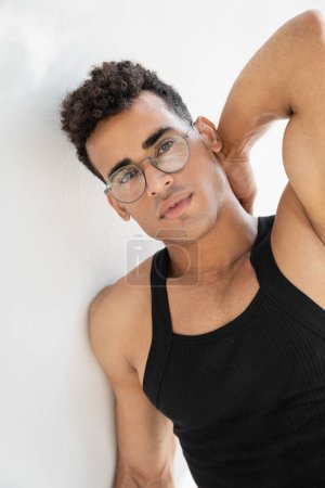 Portrait of muscular young cuban man in eyeglasses and tank top touching neck and looking at camera 
