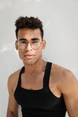 Téléchargez les photos : Portrait de jeune homme cubain bouclé dans des lunettes élégantes et t-shirt sans manches regardant la caméra - en image libre de droit