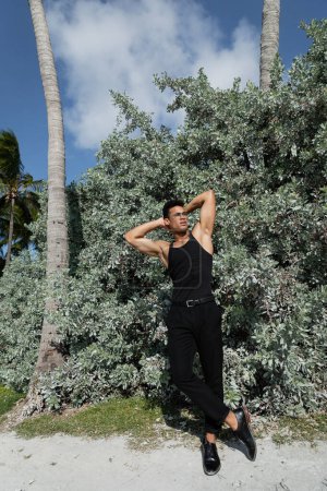 muskulöser kubanischer Mann in schwarzem Outfit und Brille in der Nähe grüner Pflanzen in Miami, Südstrand