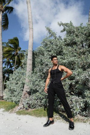 junger kubanischer Mann in schwarzem Outfit und Brille in der Nähe grüner Pflanzen im Freien in Miami, Südstrand