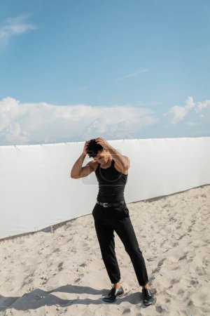 kubanischer Mann im schwarzen Outfit, der am Strand von Miami auf Sand steht und die Haare berührt 