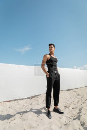 athletischer junger kubanischer Mann in schwarzem ärmellosen T-Shirt und Hose posiert in Miami, Südstrand