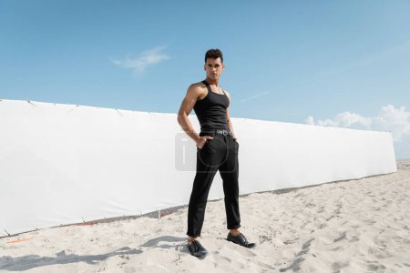 muskulöser junger kubanischer Mann in schwarzer Kleidung posiert mit Händen in Taschen in Miami, Südstrand