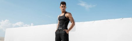 Téléchargez les photos : Jeune homme cubain musclé en tenue noire posant avec les mains dans les poches à Miami, plage sud, bannière - en image libre de droit