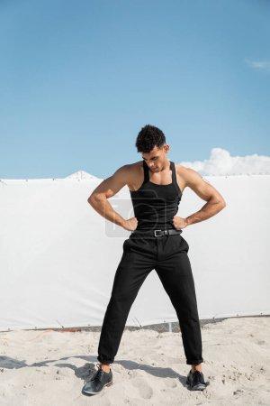 stylischer junger kubanischer Mann in schwarzem ärmellosen T-Shirt, der die Hände an den Hüften hält in Miami, Südstrand