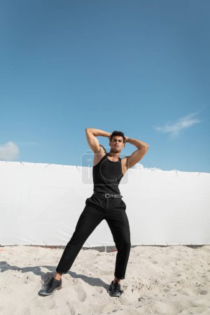 Starker und junger kubanischer Mann in schwarzem Outfit, der den Kopf berührt und in die Kamera schaut in Miami, Südstrand