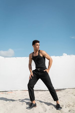 Gut aussehender kubanischer Mann in schwarzem ärmellosen T-Shirt und Hose steht auf Sand in Miami, Südstrand