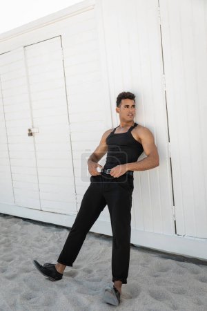 Stilvoller und muskulöser kubanischer Mann im schwarzen Outfit, der den Taillengürtel in Miami, South Beach berührt
