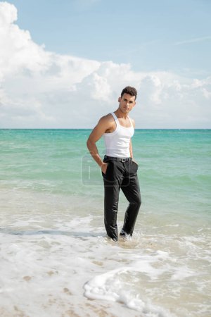 Téléchargez les photos : Jeune homme cubain musclé debout dans l'eau de l'océan à Miami South Beach, les mains dans les poches - en image libre de droit