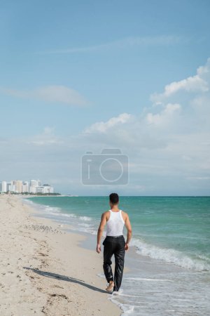 Back view of young man walking on sand near ocean water of Miami South Beach, Florida