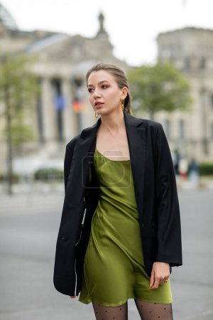 Modische und hübsche junge blonde Frau in grünem Kleid und schwarzer Jacke in Berlin
