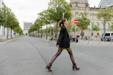 Foto de Vista lateral de la mujer joven de moda en vestido y chaqueta negra caminando por la carretera en Berlín, Alemania - Imagen libre de derechos