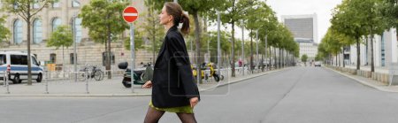 Foto de Vista lateral de la mujer joven elegante y elegante en vestido y chaqueta negra caminando en Berlín, pancarta - Imagen libre de derechos