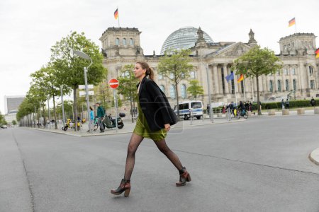 Téléchargez les photos : Pleine longueur de jeune femme blonde en robe de soie, veste et bottes marchant sur la route à Berlin - en image libre de droit