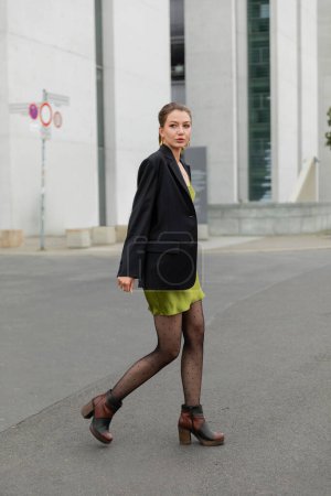 Stilvolle junge Frau in schwarzer Jacke, grünem Seidenkleid und moderner Strumpfhose in Berlin