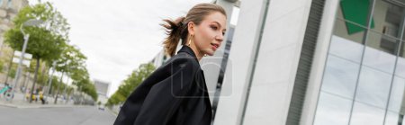 Téléchargez les photos : Jeune femme blonde positive en blazer noir regardant ailleurs tout en marchant à Berlin, bannière - en image libre de droit