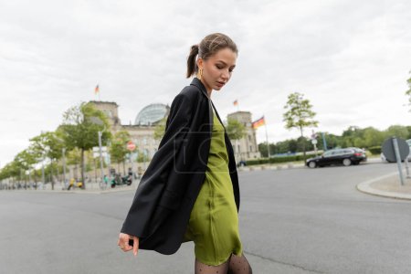 Young attractive fair haired woman in black blazer and silk dress looking down in Berlin, Germany 