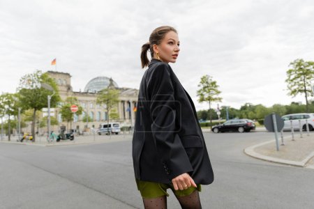 Young stylish fair haired woman in black jacket and dress looking away while walking in Berlin