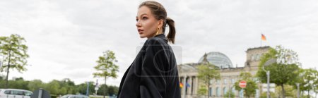 Joven mujer de pelo justo de moda en chaqueta negra mirando hacia otro lado mientras está de pie en Berlín, pancarta 