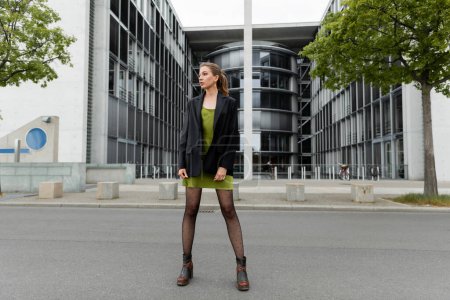 Full length of trendy young woman in boots and stylish blazer looking away  on street in Berlin