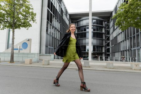 fröhliche und modische junge Frau in Blazer und grünem Seidenkleid in Berlin 