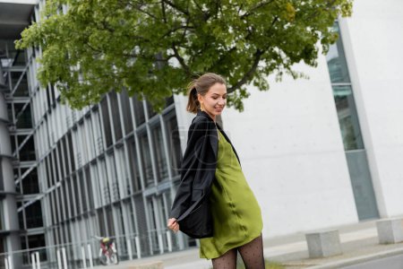 Smiling and stylish fair haired woman in silk dress and black jacket standing in Berlin, Germany