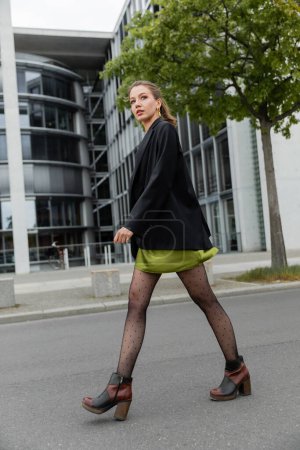 Full length of fashionable and fair haired woman in blazer, polka dot tights and boots in Berlin