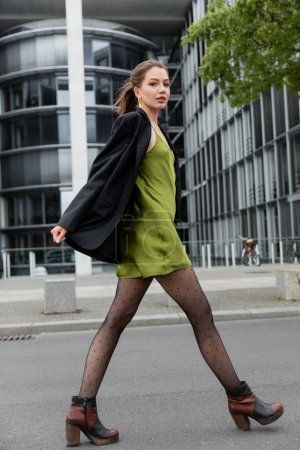 Modische junge Frau in grünem Seidenkleid, gepunkteten Strumpfhosen und Jacke blickt in Berlin in die Kamera