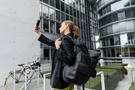 Foto de Vista lateral de la mujer joven de moda y de pelo justo con mochila tomando selfie en el teléfono inteligente - Imagen libre de derechos