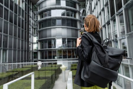 Back view of fair haired woman in black jacket holding backpack and taking photo on smartphone