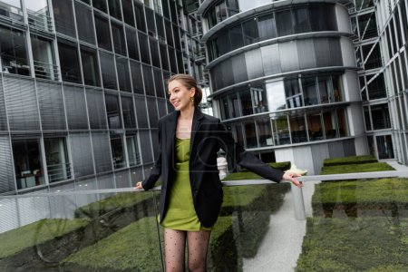 Fröhliche modische Frau in Blazer und grünem Seidenkleid schaut auf der Straße in Berlin weg 