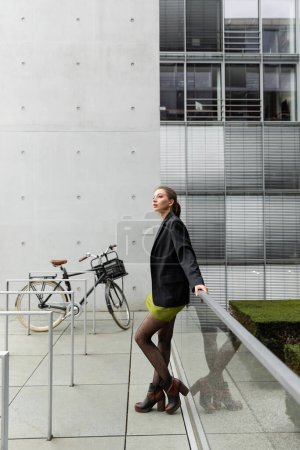 Stylische und hübsche junge Frau in schwarzer Jacke und Seidenkleid steht neben Fahrrad in Berlin 