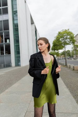 Stylish young woman in black blazer and silk dress looking away while standing in Berlin, Germany 