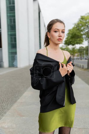 Junge blonde Frau in stylischem Seidenkleid posiert im schwarzen Blazer und blickt in Berlin in die Kamera