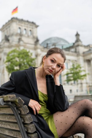 Portrait of stylish woman in jacket and silk dress looking at camera and sitting on bench in Berlin