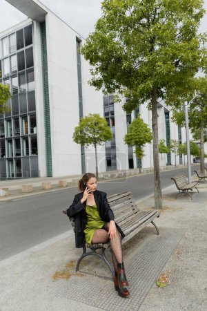 Modische Frau in Jacke, Seidenkleid und Stiefeln spricht auf dem Smartphone in Berlin 