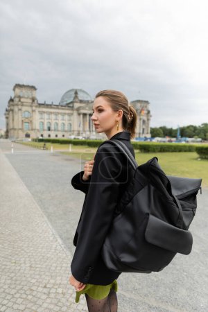 chica con chaqueta y vestido sosteniendo la mochila mientras camina cerca del edificio Reichstag en Berlín