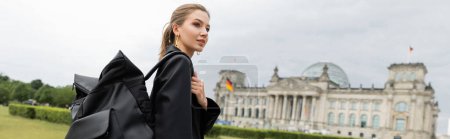 femme élégante en veste et robe tenant sac à dos tout en marchant près du bâtiment du Reichstag, bannière 