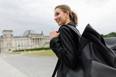 Cheerful woman in jacket and dress holding backpack while walking near Reichstag Building in Berlin
