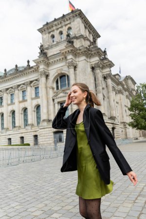 Joven alegre y elegante en blazer y vestido de seda mirando hacia otro lado cerca del edificio del Reichstag