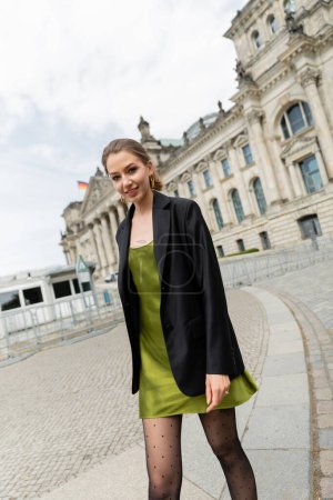 Elegante Frau in Blazer und grünem Seidenkleid beim Spaziergang in der Nähe des Reichstagsgebäudes in Berlin