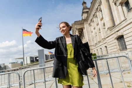 Lächelnde Frau mit Rucksack macht Selfie in der Nähe des Reichstagsgebäudes in Berlin 