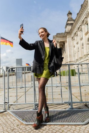 mujer alegre con mochila tomando selfie cerca del Reichstag Building en Berlín, Alemania 
