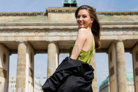 Fröhliche und hellhaarige junge Frau in Seidenkleid und Jacke blickt in die Kamera am Brandenburger Tor 