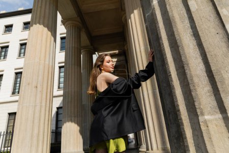 trendy young woman in silk dress holding black jacket and standing in Berlin, Germany 