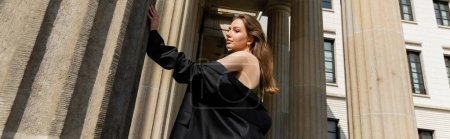 Elegant young woman in silk dress holding black jacket and standing in Berlin, banner
