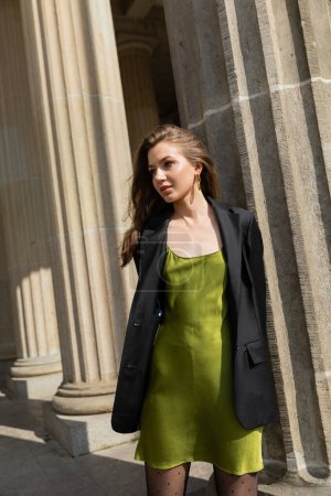 Elegant young fair haired woman in green silk dress and black jacket standing in Berlin