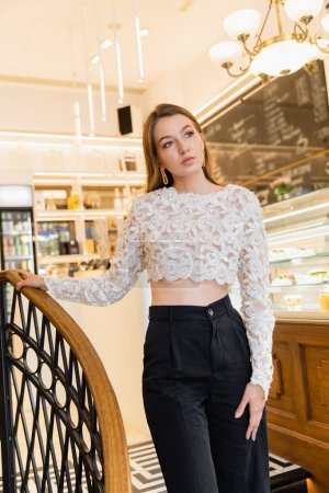 trendige und hellhaarige junge Frau in Spitzentop und Hose berührt Geländer im Café 