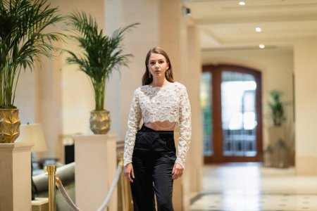 Fashionable woman in lace top and pants looking at camera while standing in modern hotel in Berlin 